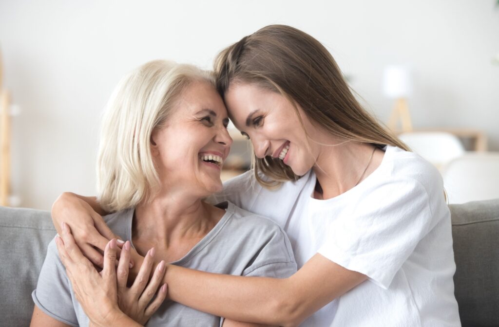 An adult child embraces their senior parent while smiling and sitting on a gray couch
