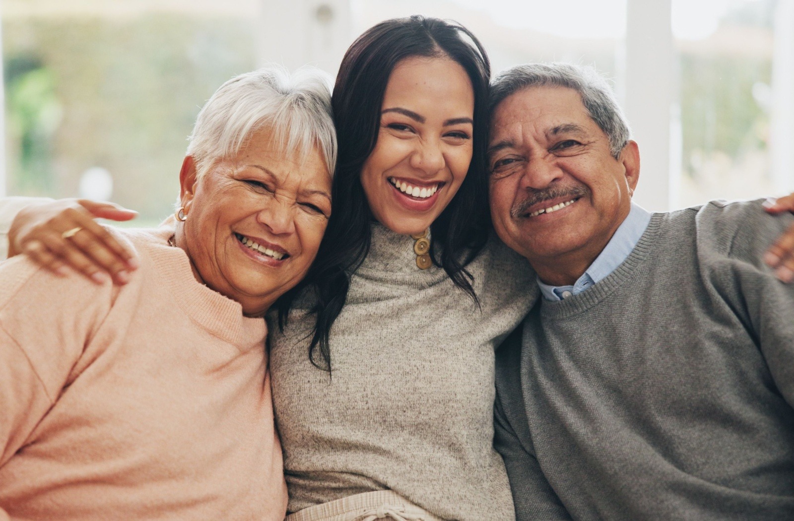 An adult child smiles as they wrap each arm around their senior parents.